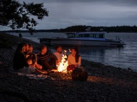 A group of friends gathers around a campfire on the beach, enjoying a cozy evening with a boat anchored nearby — perfect for memorable gatherings and sunset views.