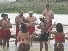 A lively group enjoys a beach celebration, capturing fun moments in the water with phones and laughter, perfect for memorable summer outings.