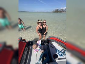 Two friends pose for a cheerful photo at the beach with a child in a life jacket nearby, capturing a joyful moment of family fun in the sun.