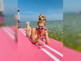 A woman enjoys a sunny day on a pink floating mat, sipping a drink and soaking up the sun — perfect for beach parties and summer fun.