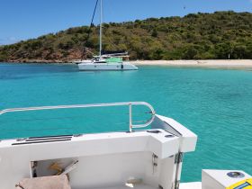A serene view of a catamaran anchored in crystal-clear turquoise waters, surrounded by lush greenery and a sandy beach — perfect for a relaxing day on the water.