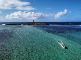 An aerial view captures a boat cruising through crystal-clear waters near a picturesque island with a lighthouse, showcasing the beauty of coastal adventures.