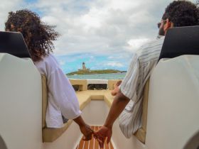 A couple enjoys a romantic moment on a boat, holding hands with a scenic lighthouse in the background — perfect for memorable outings and intimate experiences on the water.