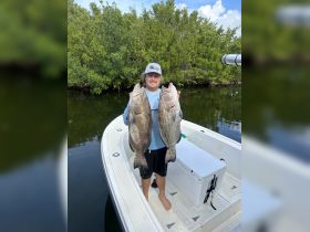 A proud angler displays his impressive catch of two large fish while aboard a boat, showcasing the thrill of fishing adventures in scenic waters.
