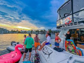 A lively group enjoys a sunset gathering on a yacht, complete with an inflatable flamingo and drinks, perfect for a fun-filled evening on the water.