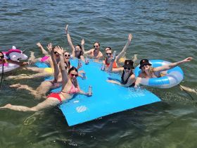 A lively group of friends enjoys a fun day on the water, relaxing on a floating mat and sipping drinks — perfect for summer celebrations and memorable outings.