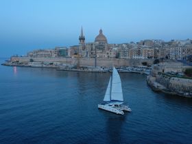 A serene aerial view of a catamaran sailing near the historic waterfront of Valletta, Malta, showcasing the beautiful architecture and calm waters.