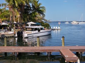 A sleek yacht is docked in a vibrant marina, surrounded by palm trees and dining areas, showcasing the perfect setting for a day on the water.