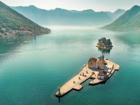 A stunning aerial view of a serene island with a historic church, surrounded by tranquil waters and majestic mountains — an idyllic destination for boat tours and sightseeing.