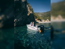 A couple enjoys a moment on a boat near a rocky coastline, with one person swimming in the crystal-clear waters — a perfect scene for a summer getaway.
