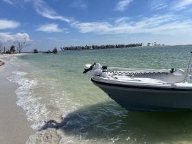 A serene view of a boat docked along a sandy shoreline, surrounded by clear waters and a picturesque backdrop of palm trees and distant buildings.