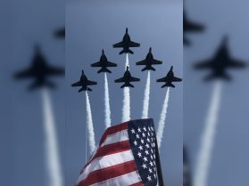 An aerial display featuring U.S. Navy jets soaring above, with an American flag in the foreground, capturing a moment of patriotic celebration.