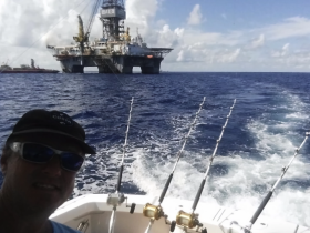 A fisherman takes a selfie aboard a boat with fishing rods in the foreground, showcasing an offshore oil rig in the background — a perfect day for deep-sea fishing adventures.