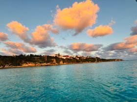 A stunning sunset view over a tranquil coastline, showcasing vibrant clouds and clear turquoise waters — perfect for capturing the beauty of nature during a boating adventure.