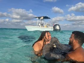 Two men interact closely with a stingray in the clear waters, showcasing a unique wildlife experience while a boat is anchored nearby.