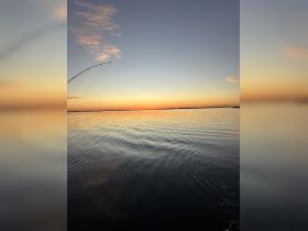 A serene sunset view over calm waters, with a fishing rod in the foreground, capturing the essence of a peaceful fishing experience.