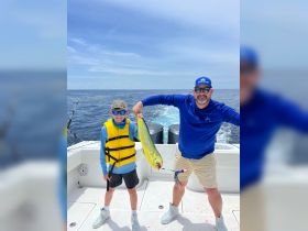 A father and son celebrate their fishing catch aboard a boat, showcasing a vibrant mahi-mahi against the backdrop of the open ocean — a perfect family fishing adventure.