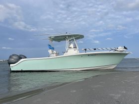 A sleek center console boat rests on the shore, showcasing its stylish design against a backdrop of blue skies and calm waters.