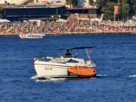 A speedboat cruises through the water, showcasing its sleek design against a backdrop of a busy beach, perfect for summer outings.