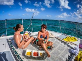 Two friends enjoy snacks and drinks on the deck of a catamaran under a sunny sky, capturing the essence of a perfect day on the water.