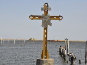 An unclear image featuring a cross near the water, with some posts visible in the foreground. The scene lacks distinct subjects or clarity.
