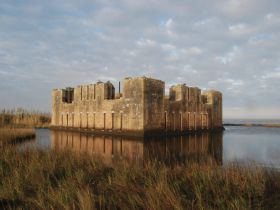 A scenic view of a historic structure reflected in calm waters, surrounded by tall grass and a cloudy sky, creating a serene atmosphere.