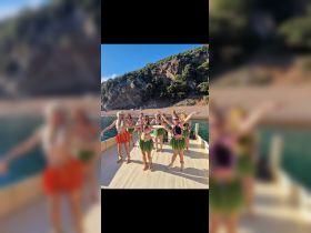 A vibrant group of dancers performs on a boat, celebrating with traditional attire and lively movements against a stunning coastal backdrop.
