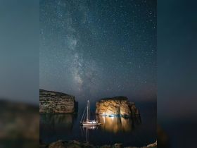 A serene night view of a sailboat anchored near dramatic cliffs under a stunning Milky Way sky, perfect for stargazing and peaceful retreats on the water.