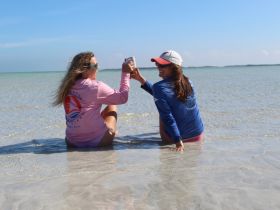 Two friends celebrate with drinks while sitting in shallow water at the beach, enjoying a sunny day — perfect for memorable moments and beach outings.