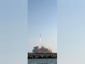 A rocket launches into the sky, creating a dramatic scene against a clear blue backdrop. The image captures the excitement of space exploration from a waterfront perspective.