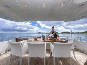 A woman prepares a beautifully arranged dining table on the deck of a yacht, showcasing a delightful spread of food and drinks against a stunning waterfront backdrop.