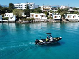 A sleek speedboat navigates through crystal-clear waters, showcasing the vibrant coastal scenery and nearby whitewashed buildings — perfect for exploring beautiful destinations.