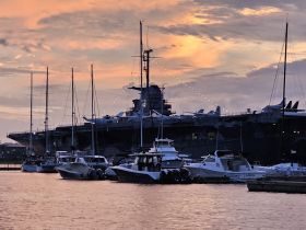 A stunning sunset view of various boats docked near a large aircraft carrier, creating a picturesque marina scene.
