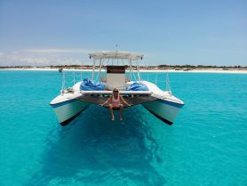 A woman relaxes on the front of a catamaran in stunning turquoise waters, with a picturesque beach in the background — an ideal setting for a tropical getaway.