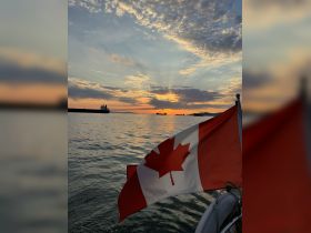 A stunning sunset view with a Canadian flag in the foreground, showcasing the serene beauty of the water and distant vessels — perfect for capturing the essence of boating in Canada.