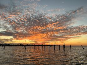 A stunning sunset paints the sky in vibrant hues over the water, with silhouettes of old pilings adding to the picturesque coastal scene.