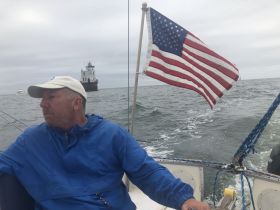 A man navigates a sailboat with an American flag waving in the wind, showcasing the serene experience of sailing on open waters.