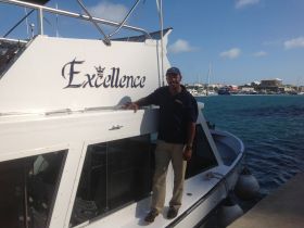 A man stands proudly by the 'Excellence' boat at the dock, showcasing the vessel's name and inviting atmosphere for potential charters.
