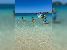 A group enjoys a fun day at the beach, wading in crystal-clear waters with a boat anchored nearby — perfect for a memorable day on the water.
