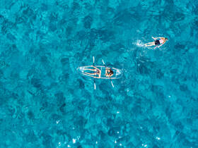 An aerial view captures two individuals enjoying a kayaking adventure in crystal-clear waters, while another swims nearby, showcasing the perfect day on the water.
