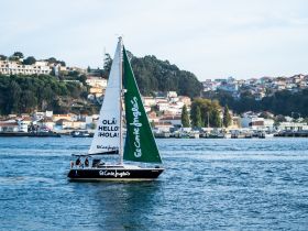 A vibrant sailboat named 'El Corte Inglés' glides through the water, showcasing its colorful sails against a picturesque coastal backdrop.