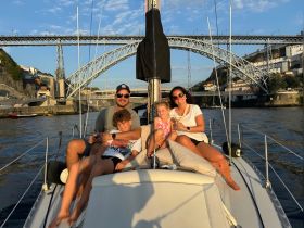 A joyful family enjoys a sunset cruise on a sailboat, capturing precious moments with drinks in hand against a stunning backdrop of a bridge.