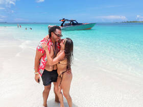 A couple shares a joyful moment on the beach, surrounded by clear turquoise waters and a boat in the background — perfect for romantic getaways and beach celebrations.