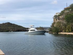 A sleek yacht navigates through calm waters, showcasing its elegant design against a scenic backdrop of hills and greenery.