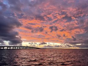 A stunning sunset paints the sky in vibrant hues over a tranquil waterway, with a bridge silhouetted against the colorful backdrop — perfect for capturing the beauty of nature.
