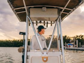 A young man at the helm of a boat, gazing towards the sunset, showcasing the serene atmosphere of a boating experience.