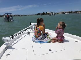 Two young girls enjoy snacks on the bow of a boat, surrounded by water and a sunny day — a perfect family outing on the water.