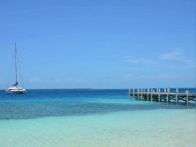 A serene view of a sailboat anchored in crystal-clear waters, with a wooden dock in the foreground — perfect for showcasing tranquil boating destinations.