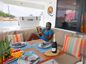 A joyful guest relaxes in the cozy interior of a yacht, enjoying a book and a glass of champagne, surrounded by a beautifully arranged dining area with fresh fruit and elegant table settings.