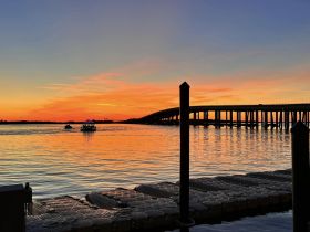 A stunning sunset over calm waters, featuring a bridge silhouette and a boat in the distance, creating a picturesque scene perfect for evening cruises.